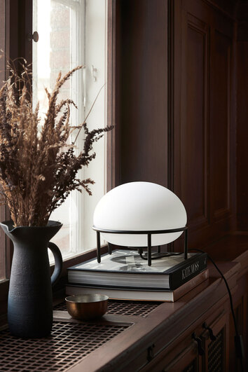 Pump Bordslampa från Woud ståendes på fönsterbrädan i ett rum med mörka träväggar.